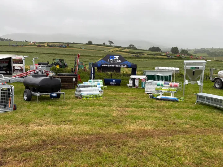 The D G Jones Agri trade stand at a grassland show - a branded gazebo with a water bowser, stacked silage wrap, hurdles, troughs and a meal bin set out on the grass.