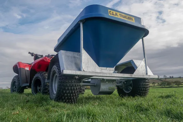 The blue hopper snacker hitched behind a quad, seen from ground level in a grass field