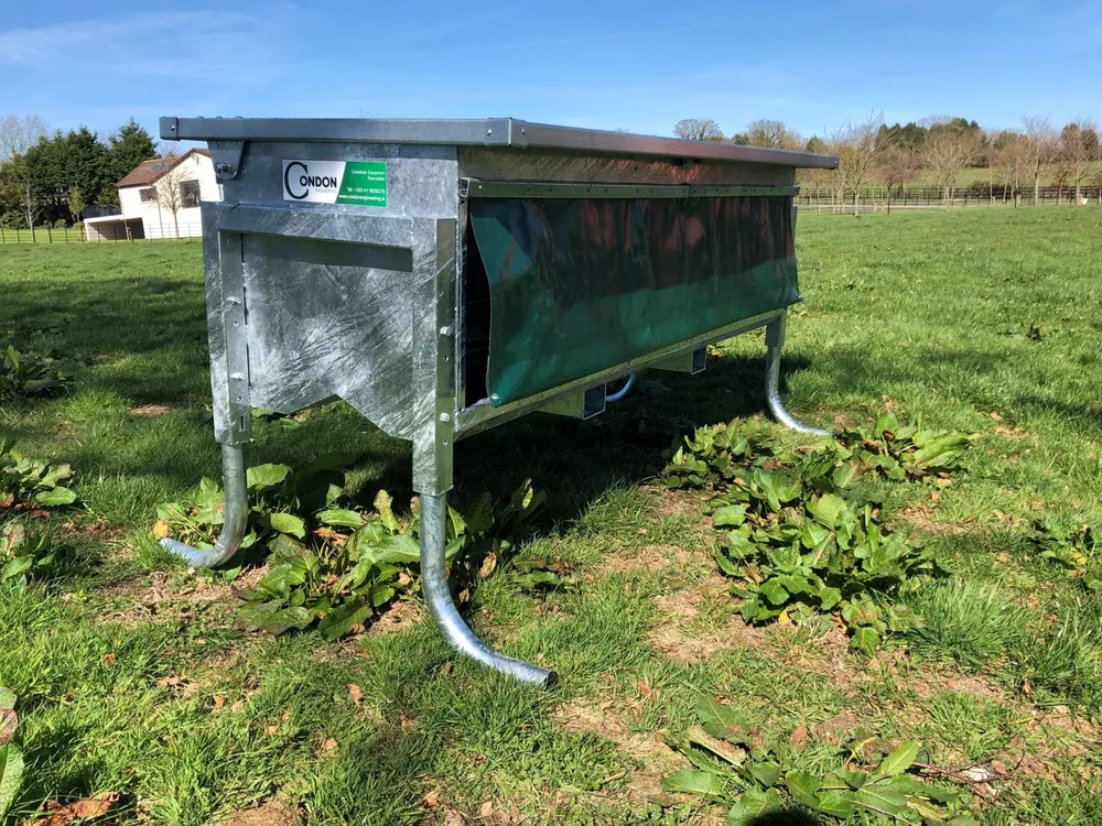 A galvanised controlled calf feeder standing on curved legs in a field, with a green rubber curtain along its open side