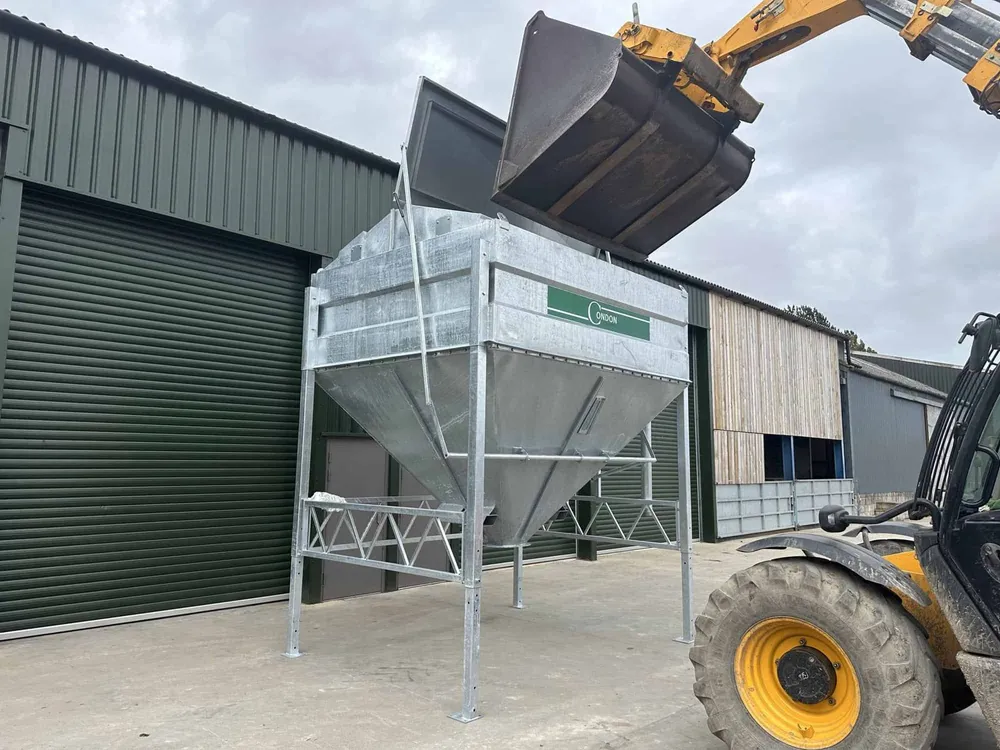 A telehandler bucket emptying into the bin through its open lid in the yard